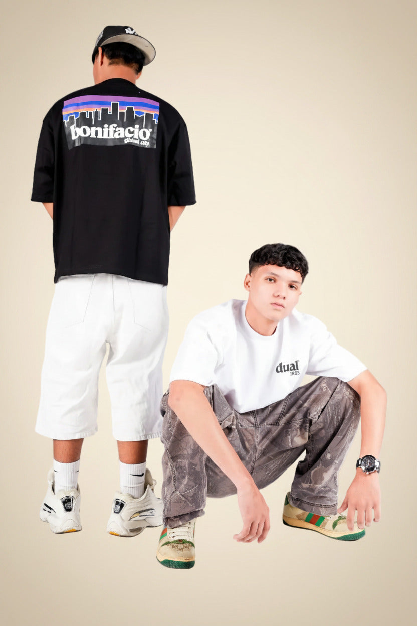 Two models in a studio setting showcasing streetwear apparel. One model stands in the back wearing a black graphic t-shirt with a colorful skyline and the brand name 'bonifacly' on the back, paired with white shorts and sneakers.